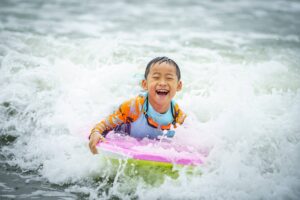 Laughing boy lying on a pink surfboard in the white water, Summer Surfkid Camp at Lower Cheung Sha