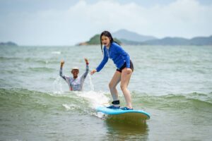 Girl in a blue rash guard surfing with a coach cheering behind her, Summer Surfkid Camp Hong Kong