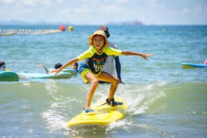 Boy in a yellow rash guard and sun hat surfing a wave, Summer Surfkid Camp Hong Kong