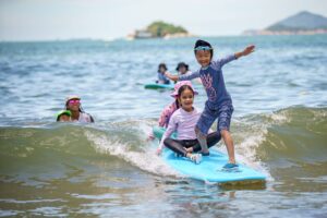 Coach steering a surfboard with a young girl standing on it, Summer Surfkid Camp Hong Kong