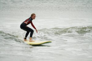 Girl in a wetsuit standing on a yellow surfboard in small waves, Easter Surfkid Camp