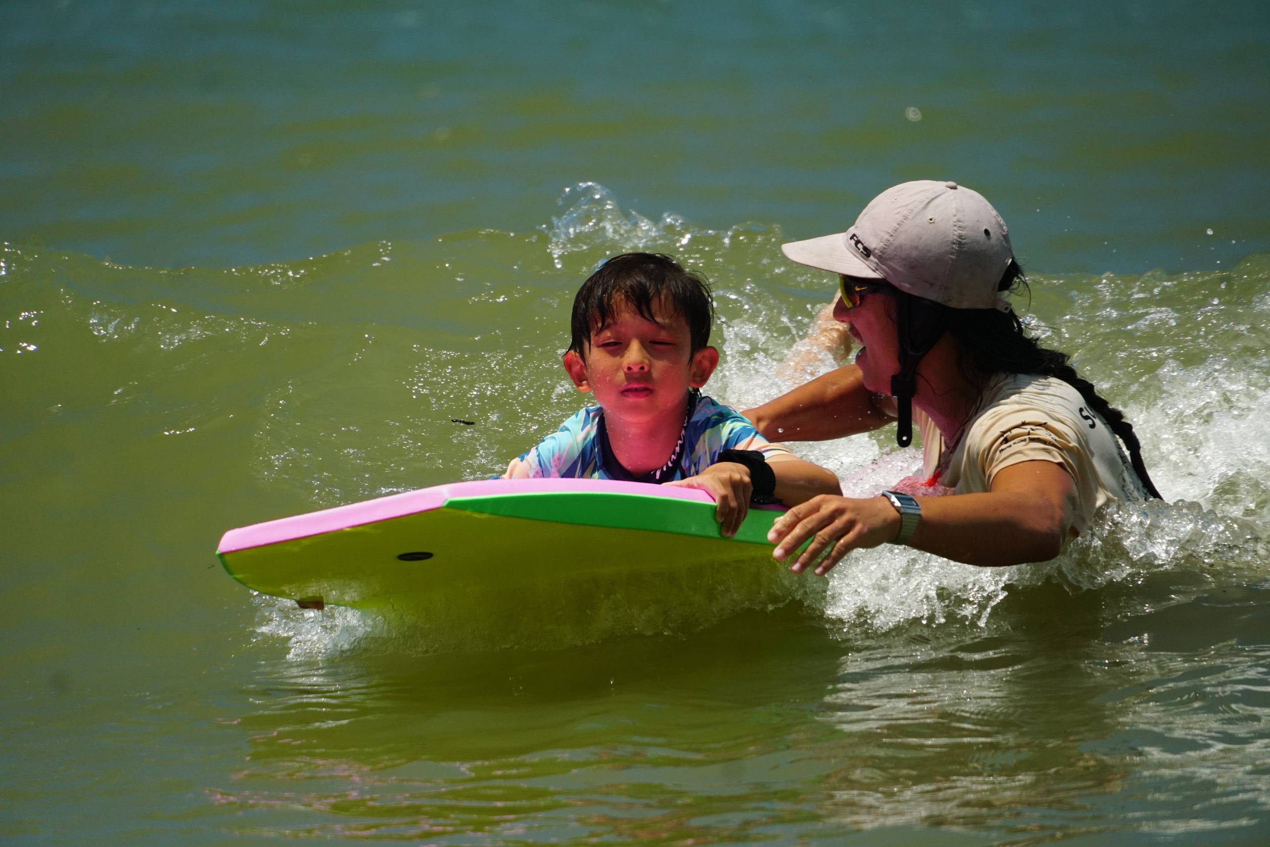 Coach guiding a young child on a surfboard in the waves at Hong Kong Surfkids Camp