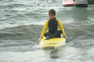 Kid in a yellow wetsuit kneeling on a surfboard in the sea, Winter Surfkid Camp Hong Kong