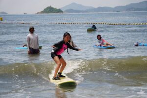Girl in a pink rash guard surfing with the Lantau hills behind, Autumn Surfkid Camp at Lower Cheung Sha
